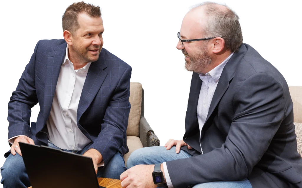 Two men in business attire sit on a couch, engaged in a lively discussion about Infrastructure Cost Estimating, with one holding a laptop and both smiling.