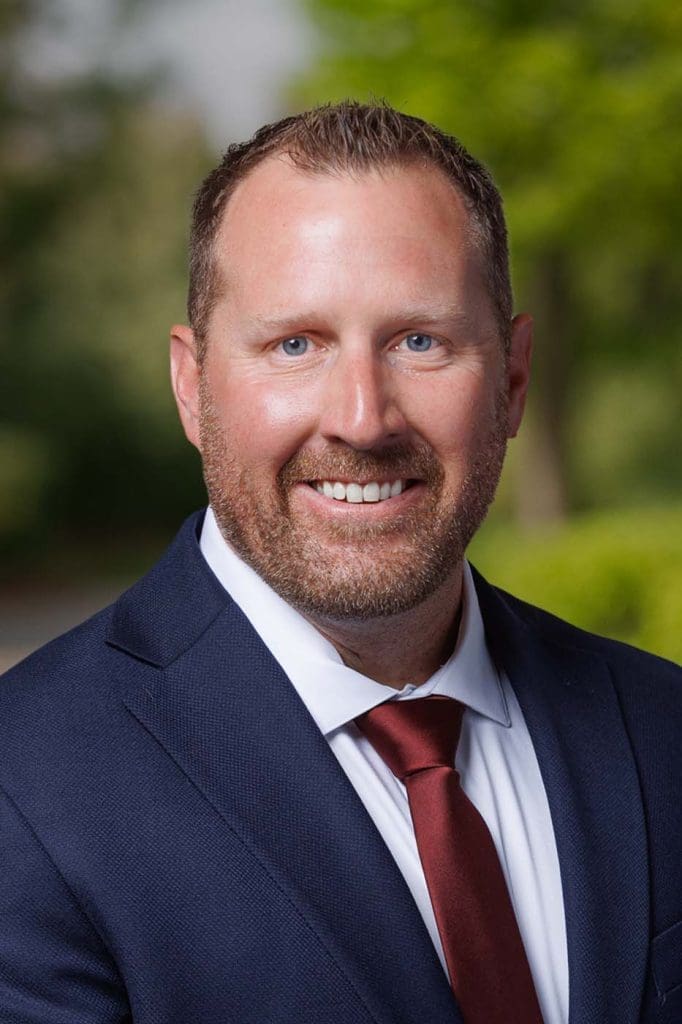Man in a blue suit, white shirt, and red tie smiling outdoors with greenery in the blurred background—perfectly embodying the confidence and professionalism valued by Independent Cost Estimators.