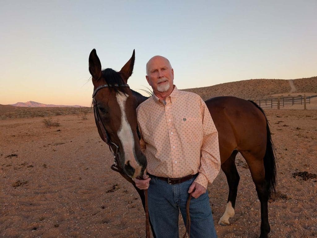 An older man stands beside a brown horse on a dirt path in a desert landscape at sunset.