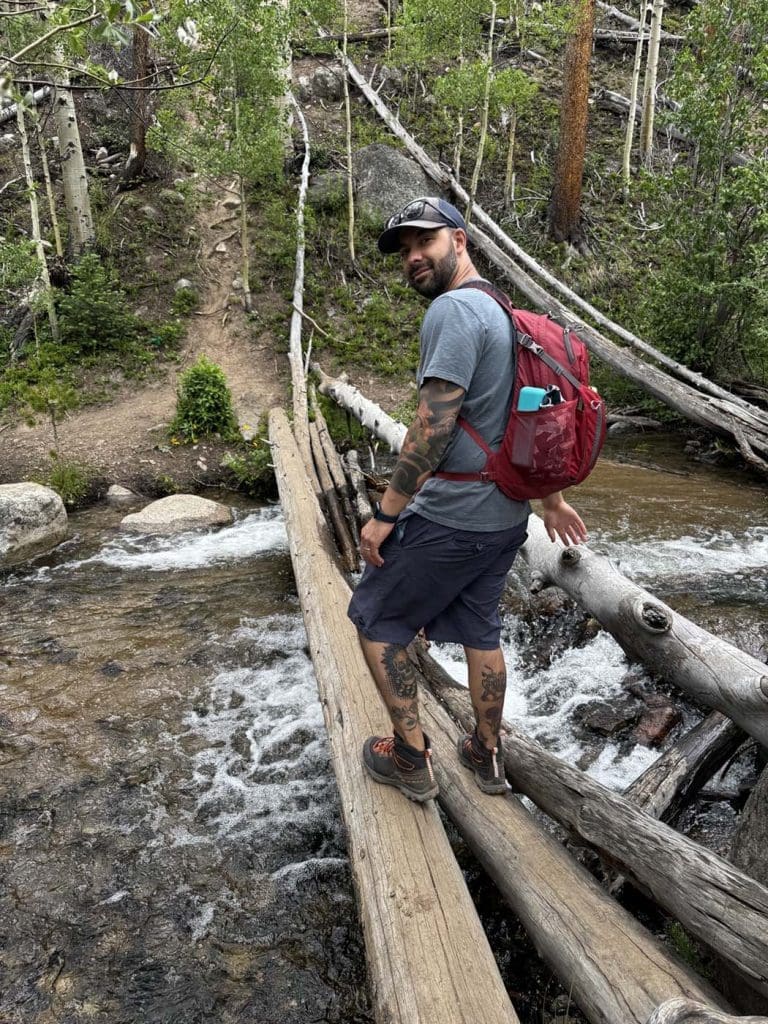 A man with a red backpack and tattoos crosses a stream on a makeshift log bridge in a forested area.
