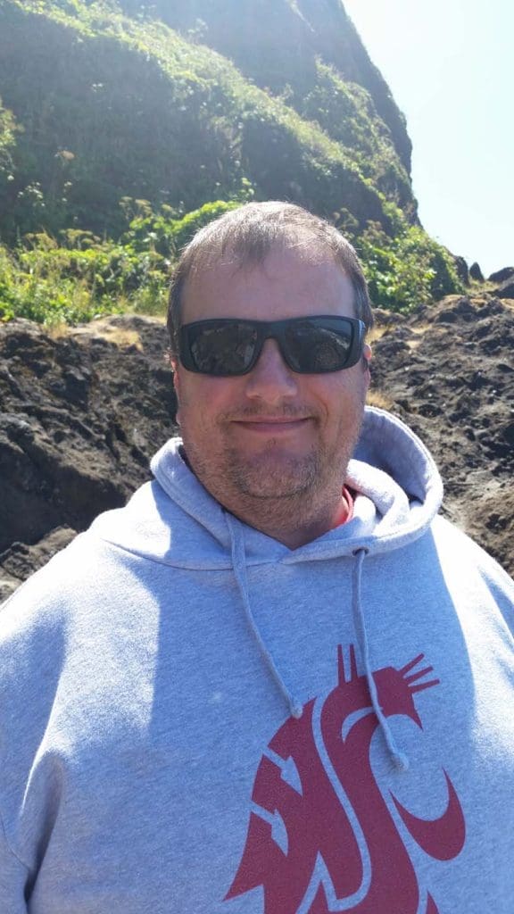A man wearing dark sunglasses and a gray hoodie with a red logo stands outdoors in front of rocky terrain and green foliage.