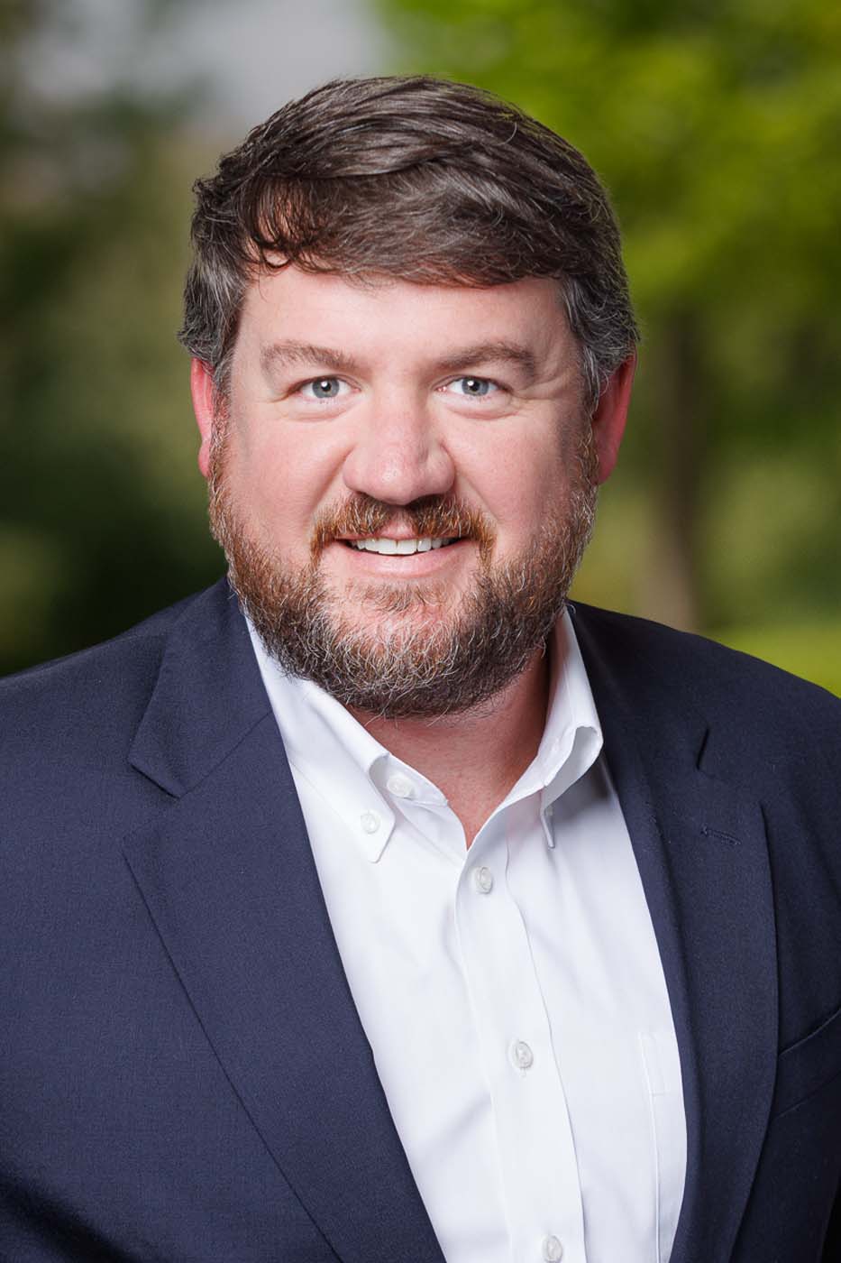 A man with short brown hair and a beard, wearing a navy blazer and white shirt, smiles at the camera with a blurred green outdoor background—reflecting the confidence of experienced Independent Cost Estimators.