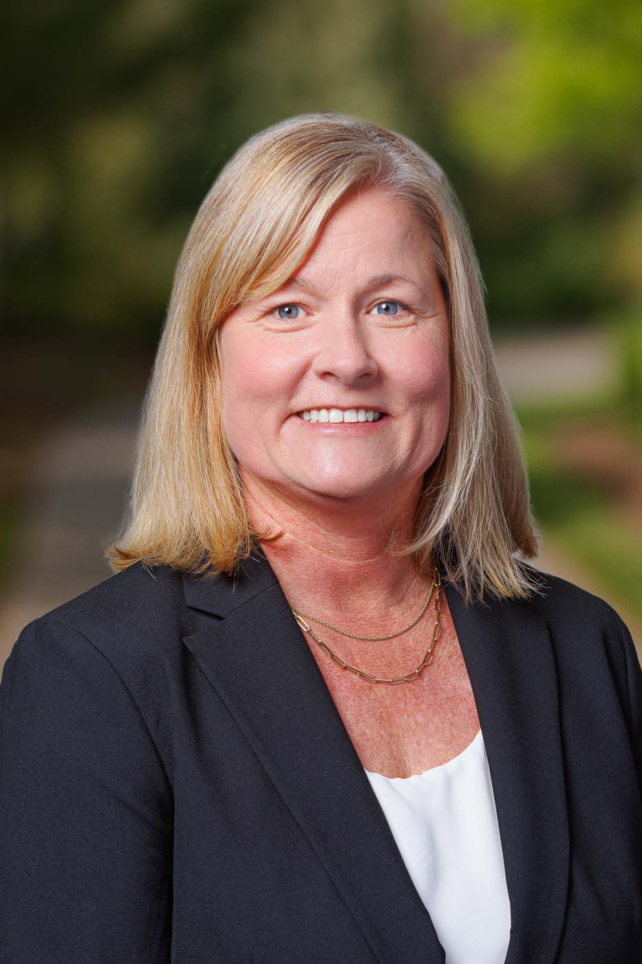 A woman with blonde hair, wearing a black blazer, white blouse, and gold necklace, smiles outdoors with greenery in the background—reflecting the confidence and professionalism of Independent Cost Estimators.