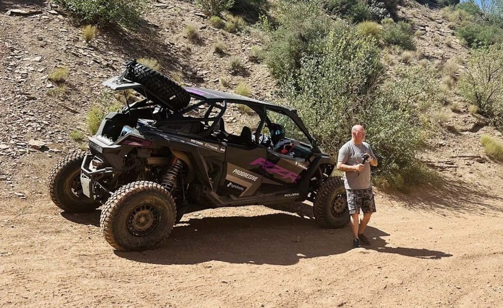A man stands next to a parked off-road vehicle on a dirt trail surrounded by rocky and grassy desert terrain.