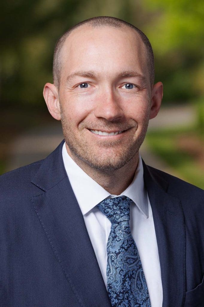 A man in a navy suit and patterned tie smiles at the camera, standing outdoors with a blurred green background—reflecting the professionalism of Independent Cost Estimators.