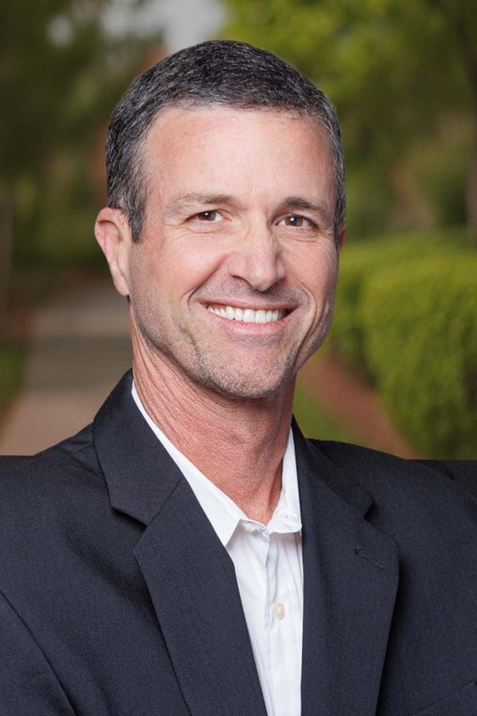 A man in a dark suit jacket and white shirt, smiling confidently, stands outdoors with greenery and a pathway in the background—capturing the professionalism often seen among Independent Cost Estimators.