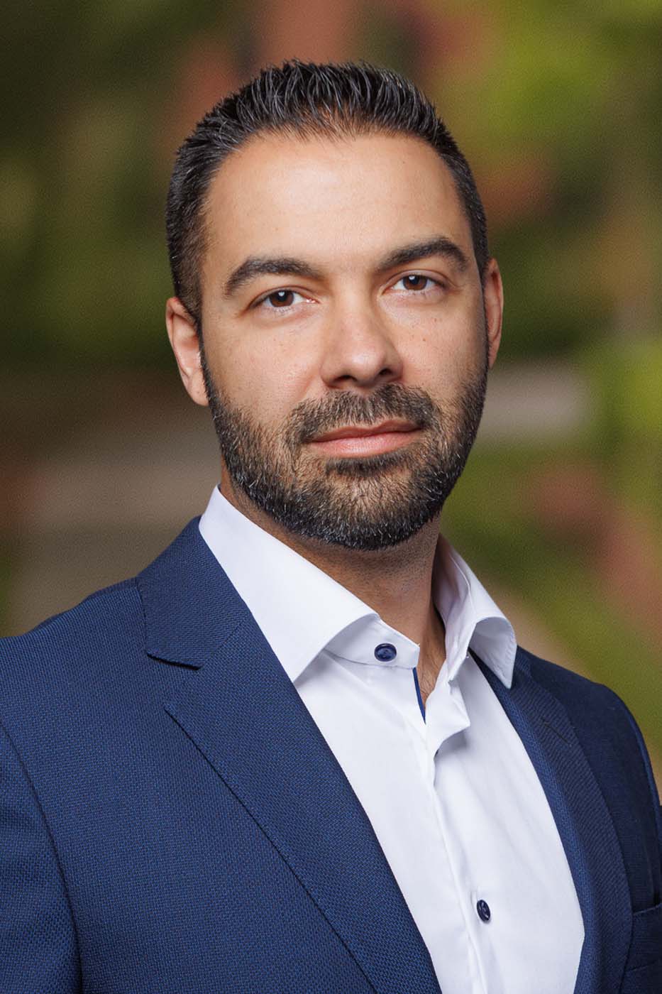 A man with short dark hair and a trimmed beard, wearing a blue suit jacket and white dress shirt, poses confidently—reflecting the professionalism of Independent Cost Estimators—against a blurred outdoor background.