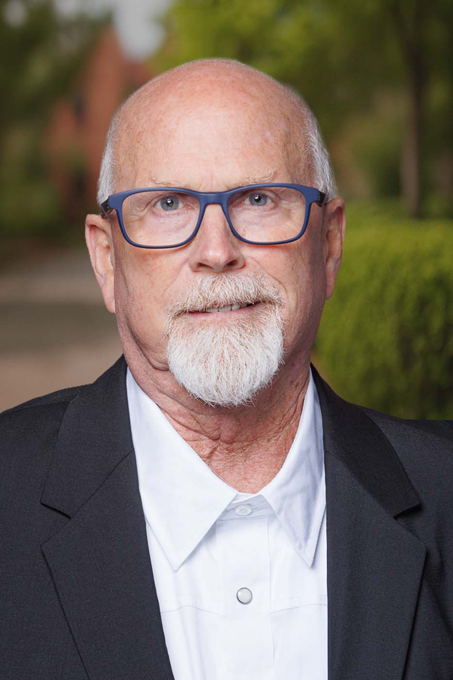 An older man with a white beard and mustache, wearing glasses, a black suit jacket, and a white shirt stands outdoors in front of greenery, embodying the professionalism often seen in experienced Independent Cost Estimators.