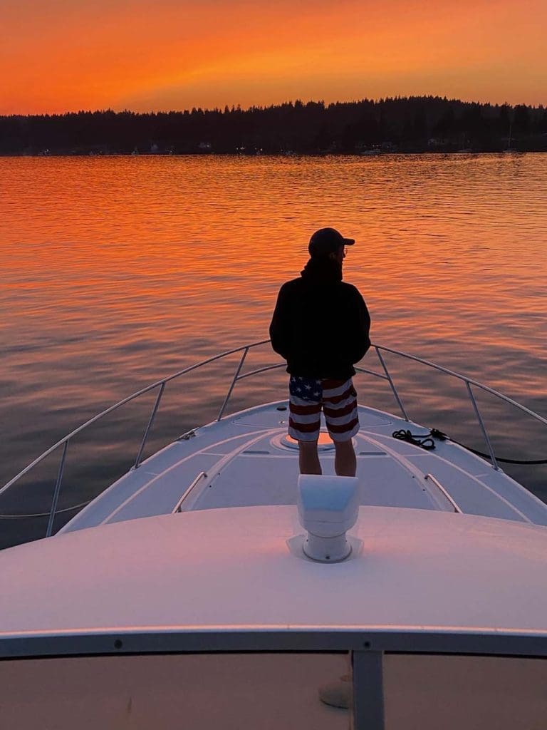 A person in a cap and American flag shorts stands on the bow of a boat, facing a calm lake during an orange sunset.