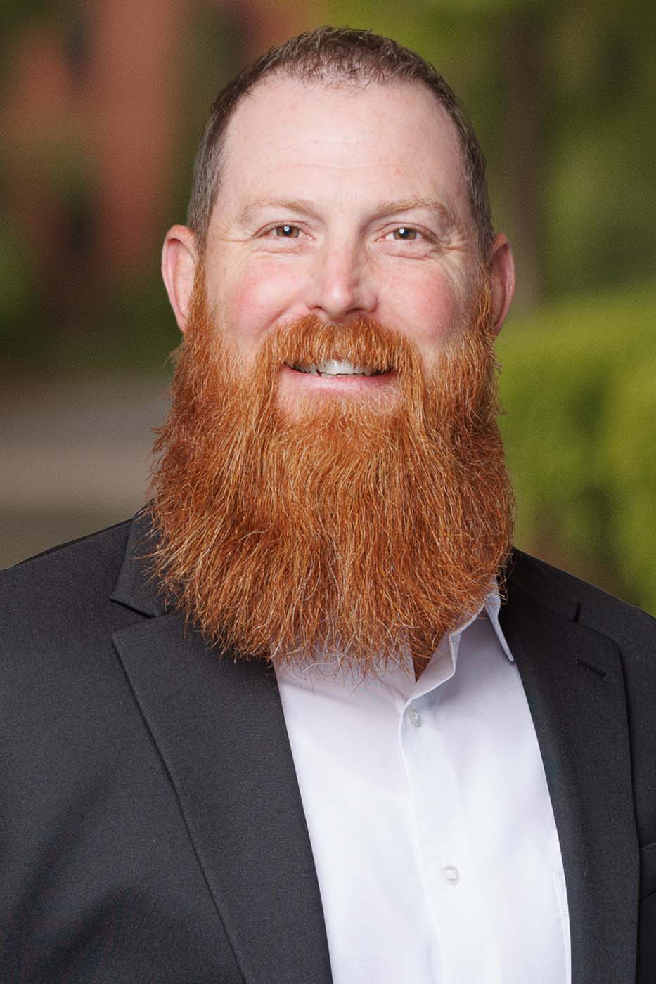 Man with a large red beard and short hair, wearing a black suit jacket and white shirt, smiling outdoors with a blurred green background—professional and approachable, ideal for roles such as Independent Cost Estimators.