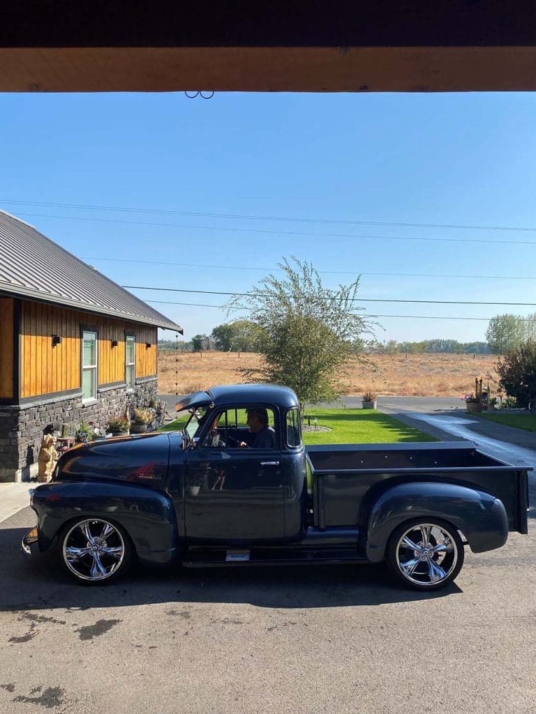 A classic black pickup truck with chrome wheels parked in a driveway beside a house on a sunny day.