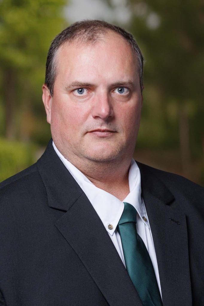 A man in a black suit, white shirt, and green tie—representing Independent Cost Estimators—looks directly at the camera against an outdoor background with greenery.