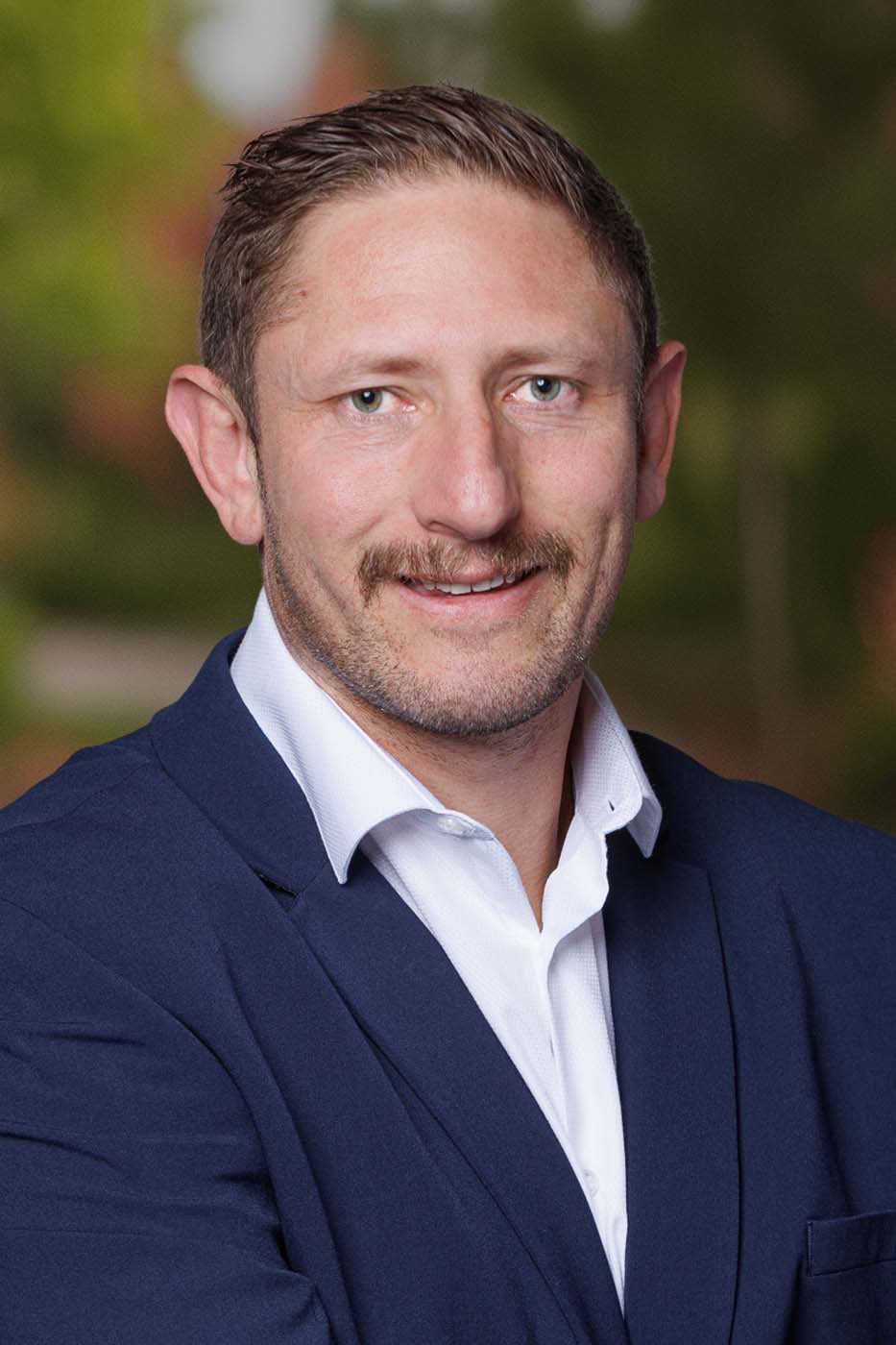 A man in a navy blazer and white shirt, representing Independent Cost Estimators, is smiling at the camera with a blurred outdoor background.