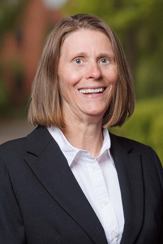 A woman with straight, shoulder-length light brown hair—one of our skilled Independent Cost Estimators—is wearing a black blazer and white shirt, smiling outdoors with greenery and a blurred building in the background.