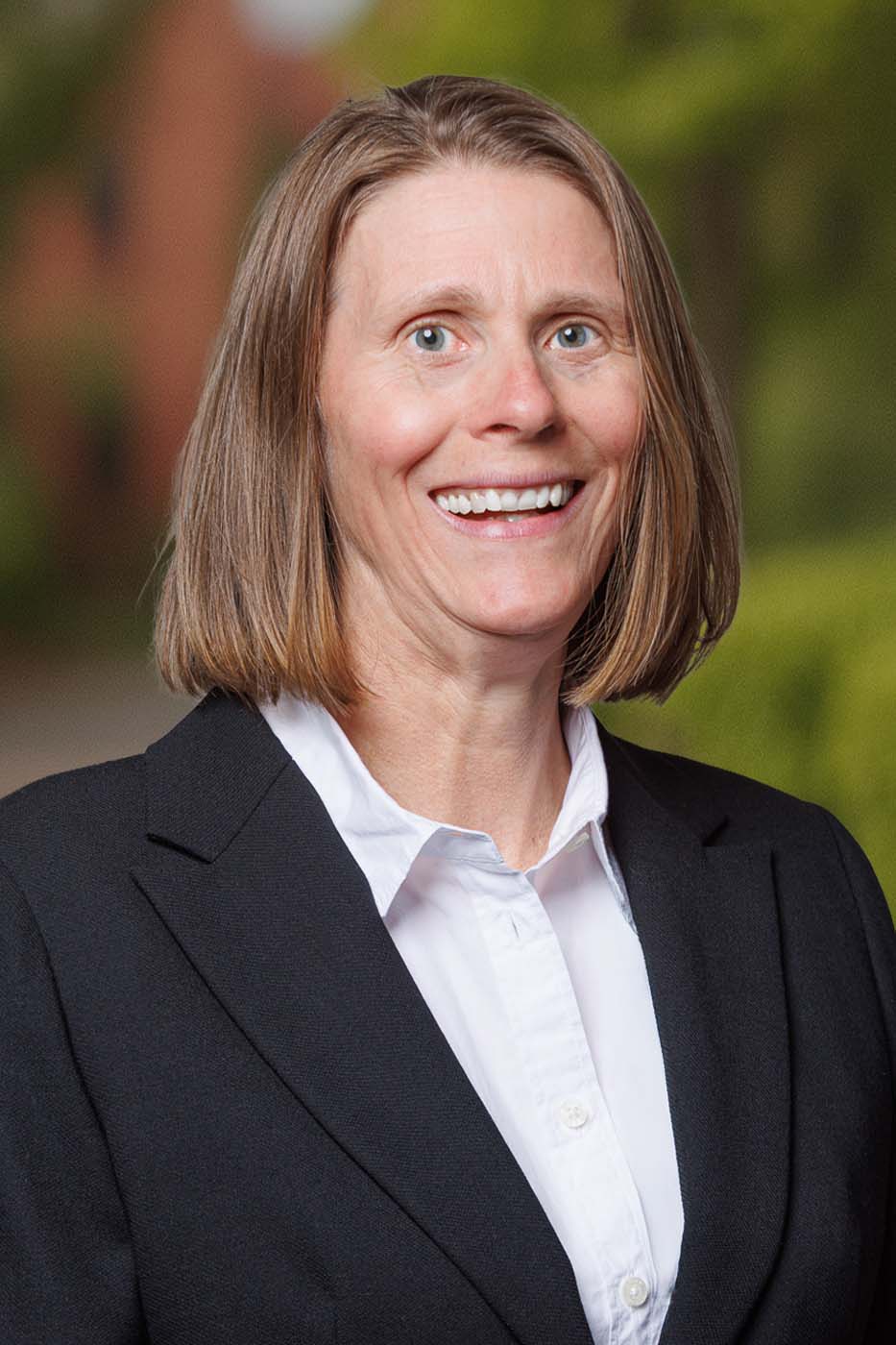 A woman with straight, shoulder-length light brown hair—one of our skilled Independent Cost Estimators—is wearing a black blazer and white shirt, smiling outdoors with greenery and a blurred building in the background.