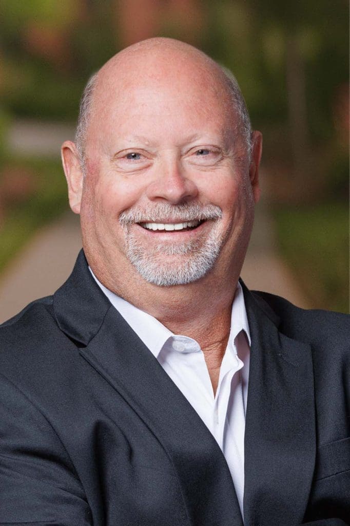 A bald man with a white goatee is smiling, wearing a black suit jacket and white shirt, standing outdoors with a blurred green background—he exudes the confidence of seasoned Independent Cost Estimators.