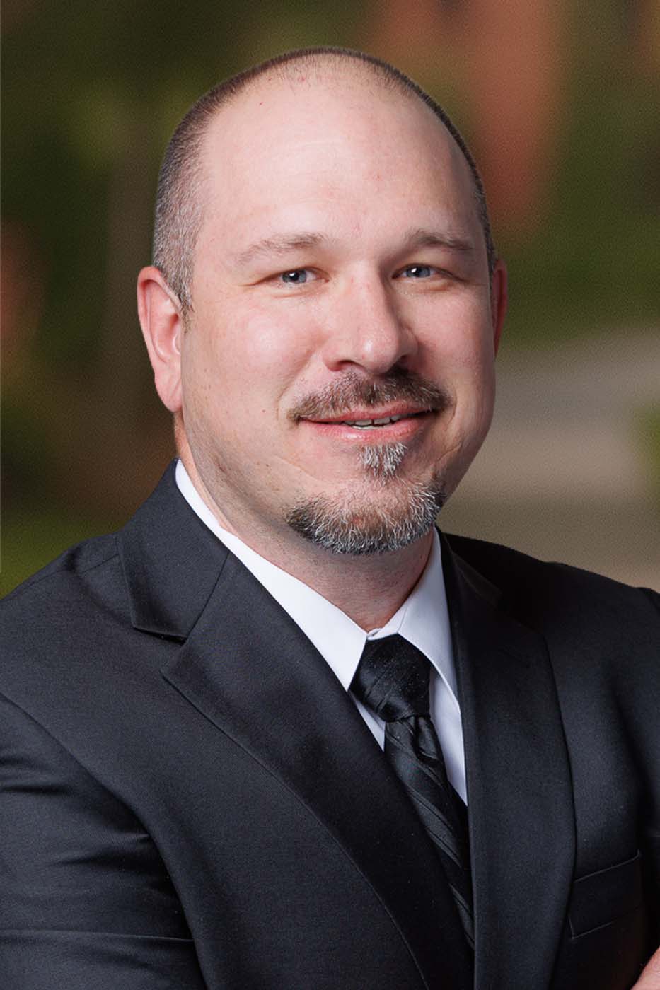 Man with short hair and a goatee, dressed in a black suit, white shirt, and black tie, smiles slightly in front of a blurred outdoor background, projecting the professionalism often seen among Independent Cost Estimators.
