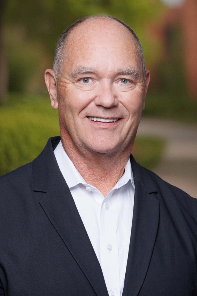 A middle-aged man with short hair, wearing a white shirt and dark blazer, smiles at the camera outdoors with greenery in the background—confident and approachable, he represents experienced Independent Cost Estimators.