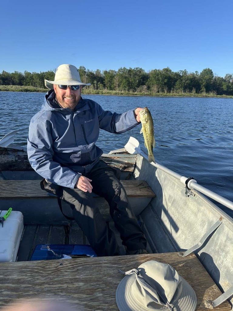 A person in a gray jacket and sun hat sits in a boat on a lake, holding up a fish, with trees visible in the background under a clear blue sky.