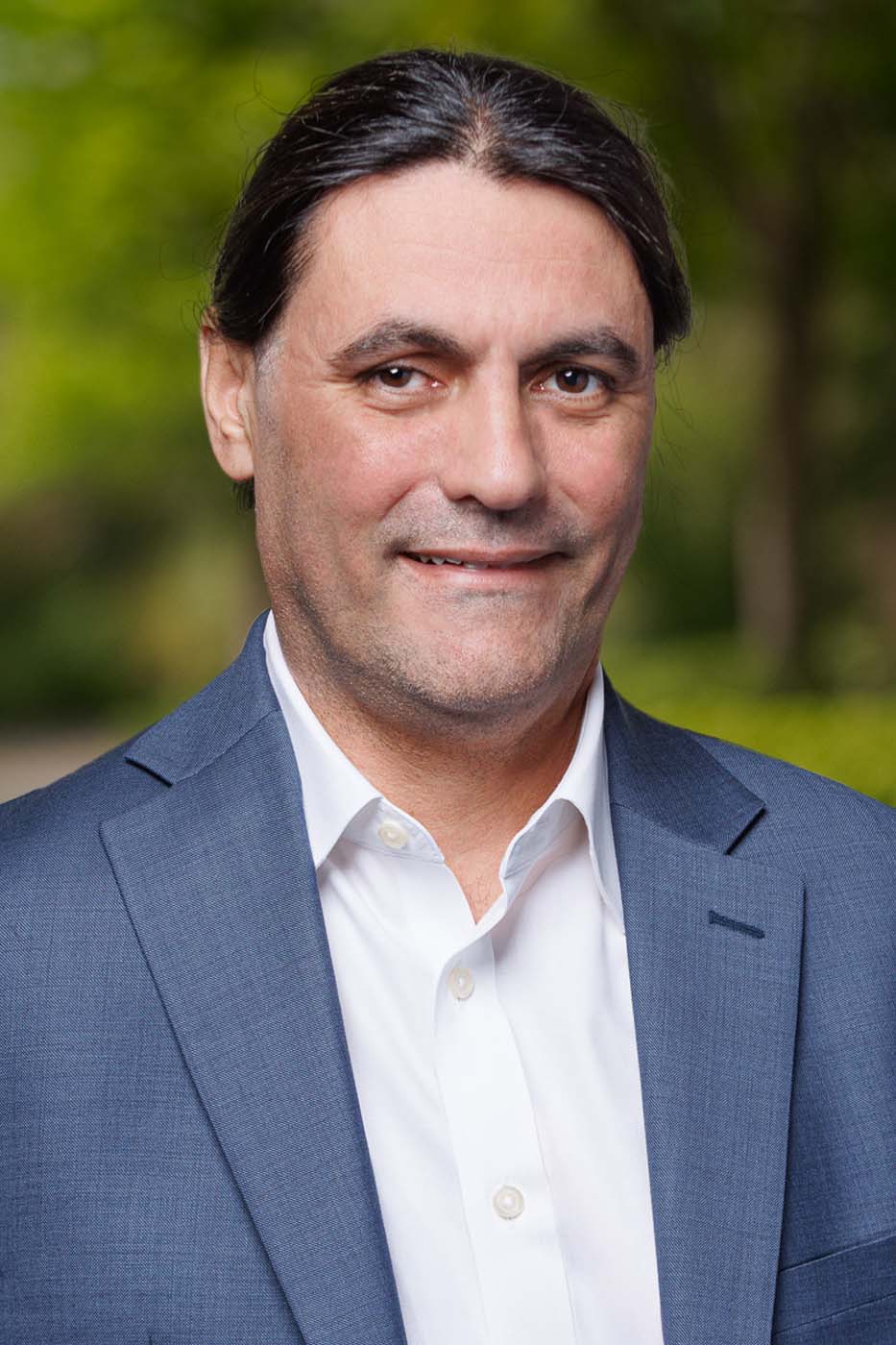 Man with long dark hair wearing a blue suit jacket and white shirt, standing outdoors with greenery in the background—an approachable look often favored by Independent Cost Estimators in professional settings.