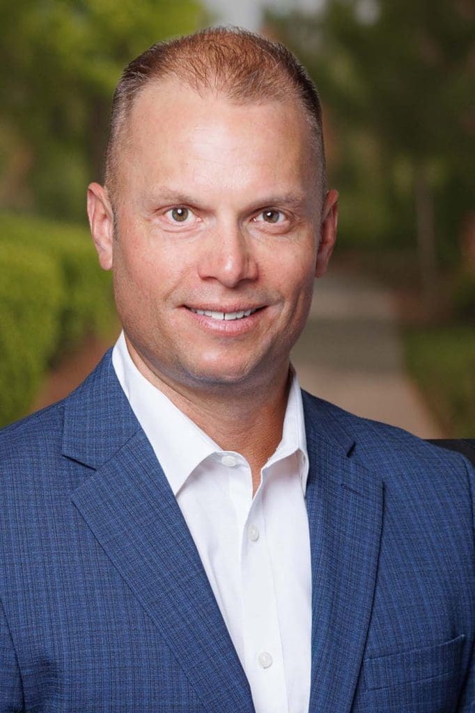 A man in a blue suit and white shirt, possibly an Independent Cost Estimator, is smiling at the camera with greenery and a pathway blurred in the background.