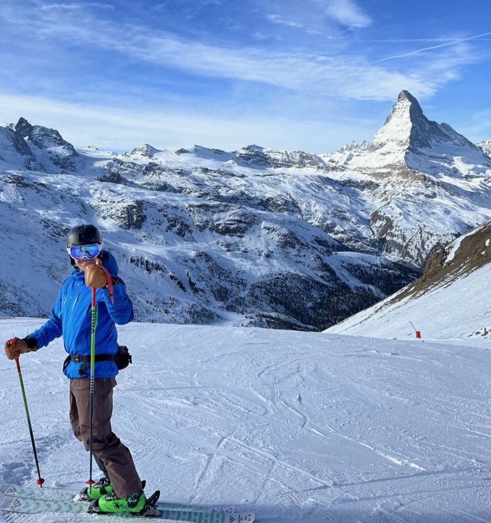 A skier stands on a snowy slope with ski poles, wearing blue jacket and helmet, with the Matterhorn mountain and snowy peaks in the background.