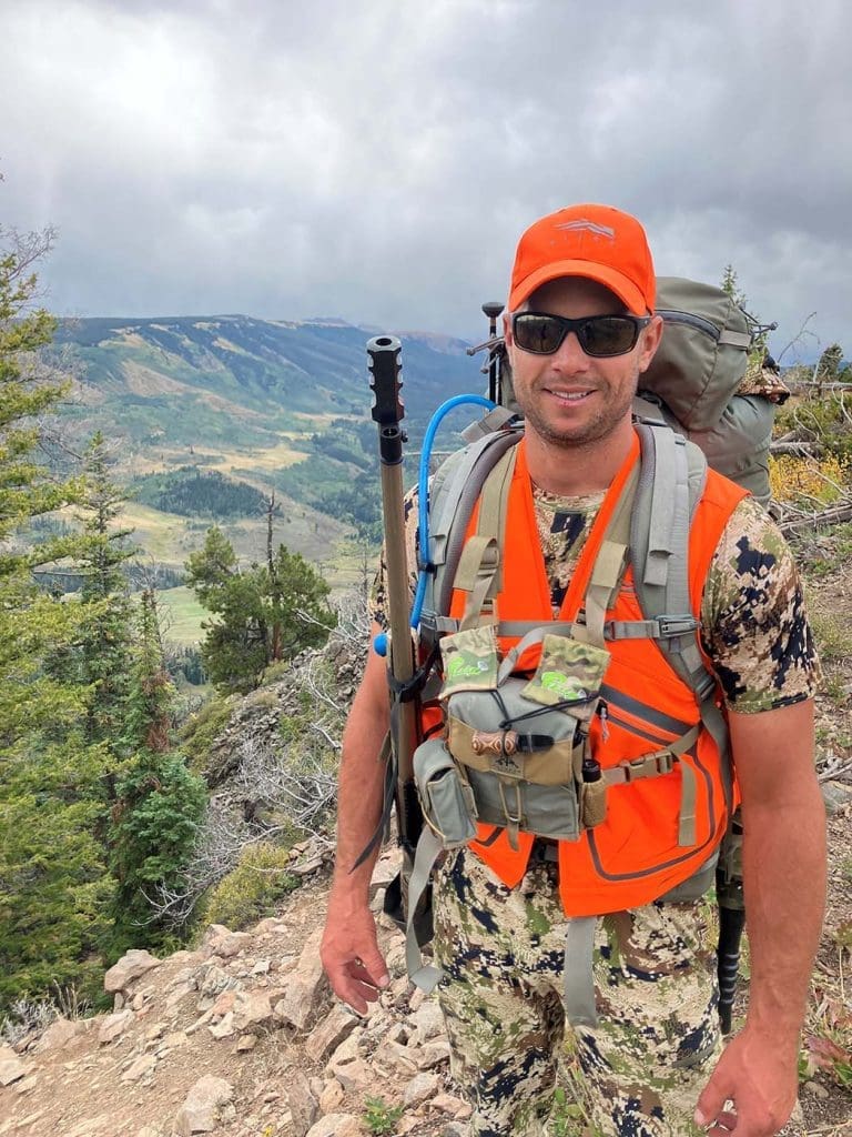 Man wearing camouflage hunting gear and orange vest and cap stands on a rocky mountain trail with a backpack, trekking pole, and scenic valley in the background.