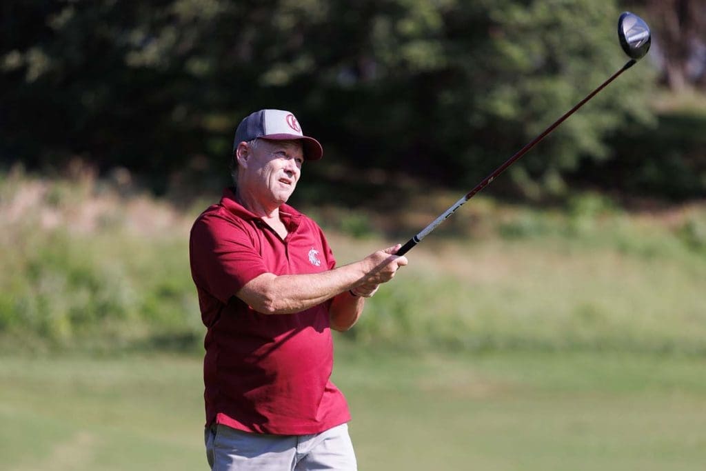 A man in a red polo shirt and cap holds a golf club and looks into the distance on a golf course.