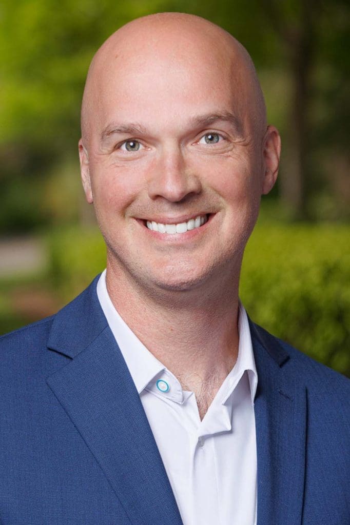 A bald man in a blue suit jacket and white shirt, representing Independent Cost Estimators, smiles at the camera outdoors, with greenery in the blurred background.