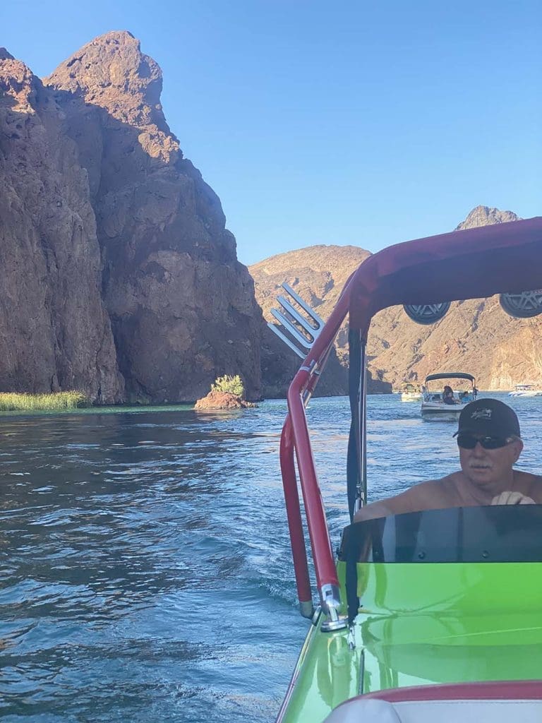 A man is driving a green and red speedboat on a river surrounded by rocky cliffs, with another boat visible in the background under a clear blue sky.