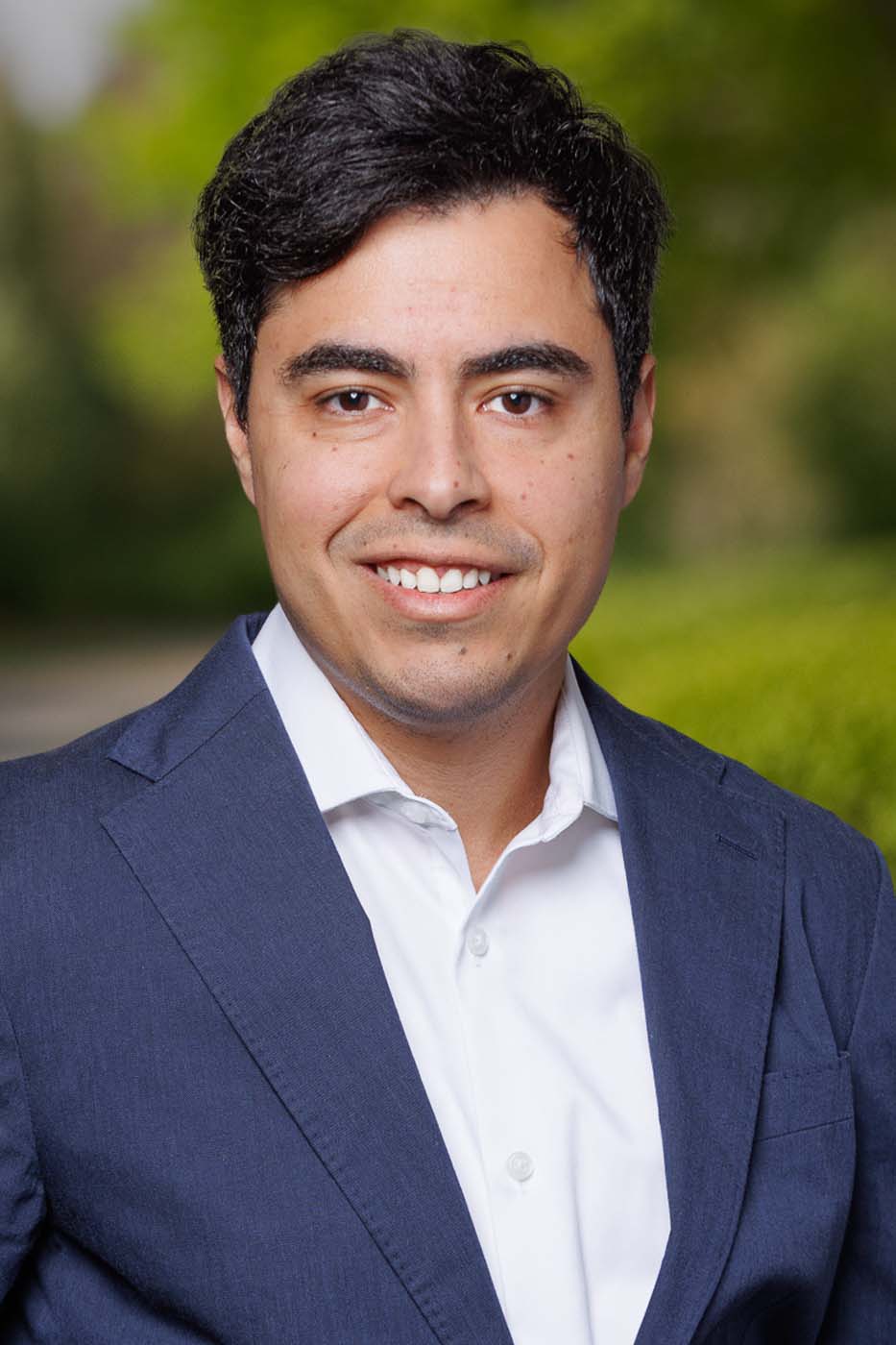 A man in a navy blue suit and white shirt smiles at the camera outdoors, with greenery and a blurred background behind him—exuding the professionalism often seen among Independent Cost Estimators.