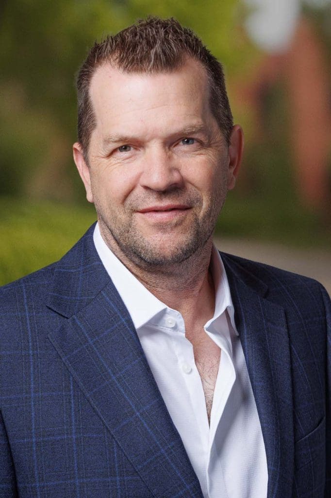 A middle-aged man with short brown hair and a trimmed beard, wearing a navy checkered blazer over a white shirt, stands outdoors with greenery in the background, embodying the professionalism of Independent Cost Estimators.