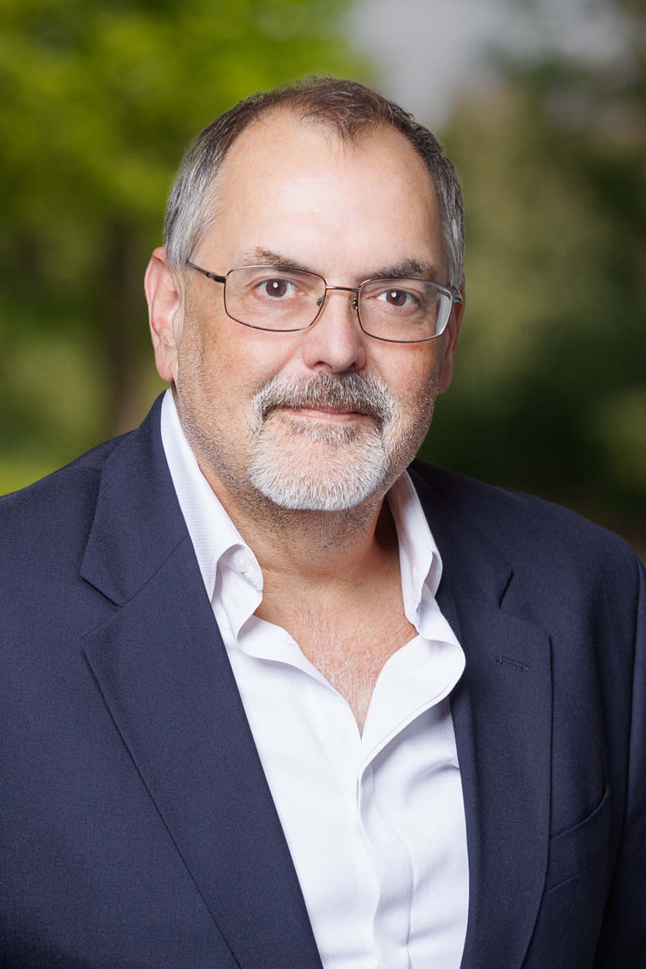 A middle-aged man with glasses, gray hair, and a beard wears a dark suit jacket over an open-collared white shirt, standing outdoors with a blurred green background—capturing the professional presence of Independent Cost Estimators.