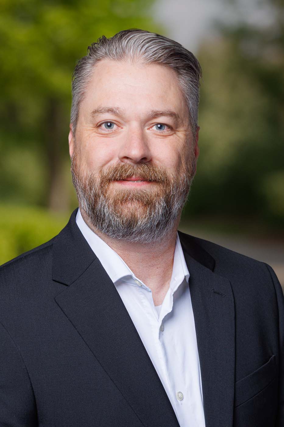 A man with gray hair and a beard, wearing a dark suit jacket and white shirt, poses outdoors with greenery in the background—reflecting the professionalism of Independent Cost Estimators.