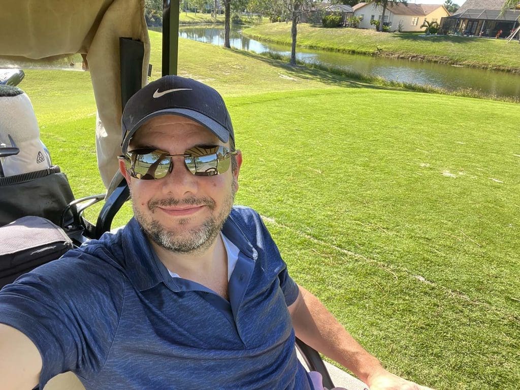 Man in sunglasses and a cap takes a selfie while sitting in a golf cart on a sunny day, with a green golf course and a pond in the background.