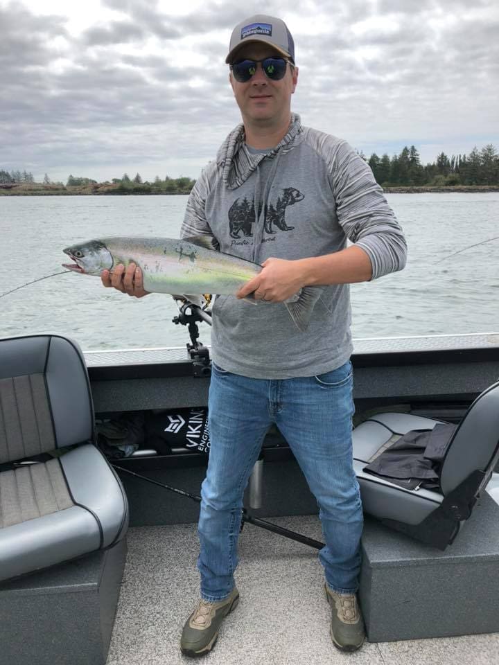 A person standing on a boat holds a fish with both hands; fishing gear and benches are visible, and water with trees is in the background.