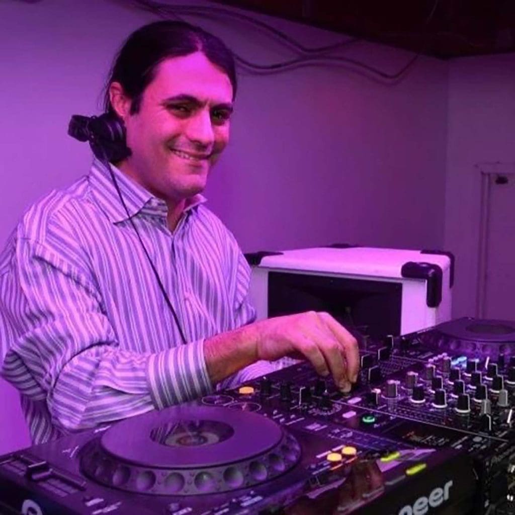 A man with long dark hair operates a DJ mixer and turntable, smiling at the camera in a room with purple lighting.