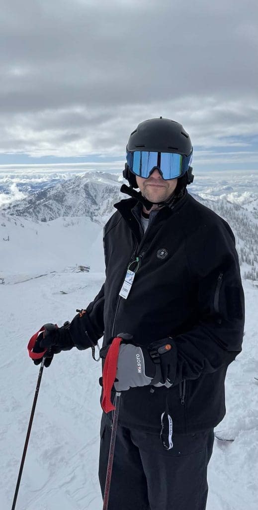 A skier wearing a black helmet, goggles, and jacket stands on a snowy mountain slope holding ski poles, with snow-covered peaks in the background under a cloudy sky.