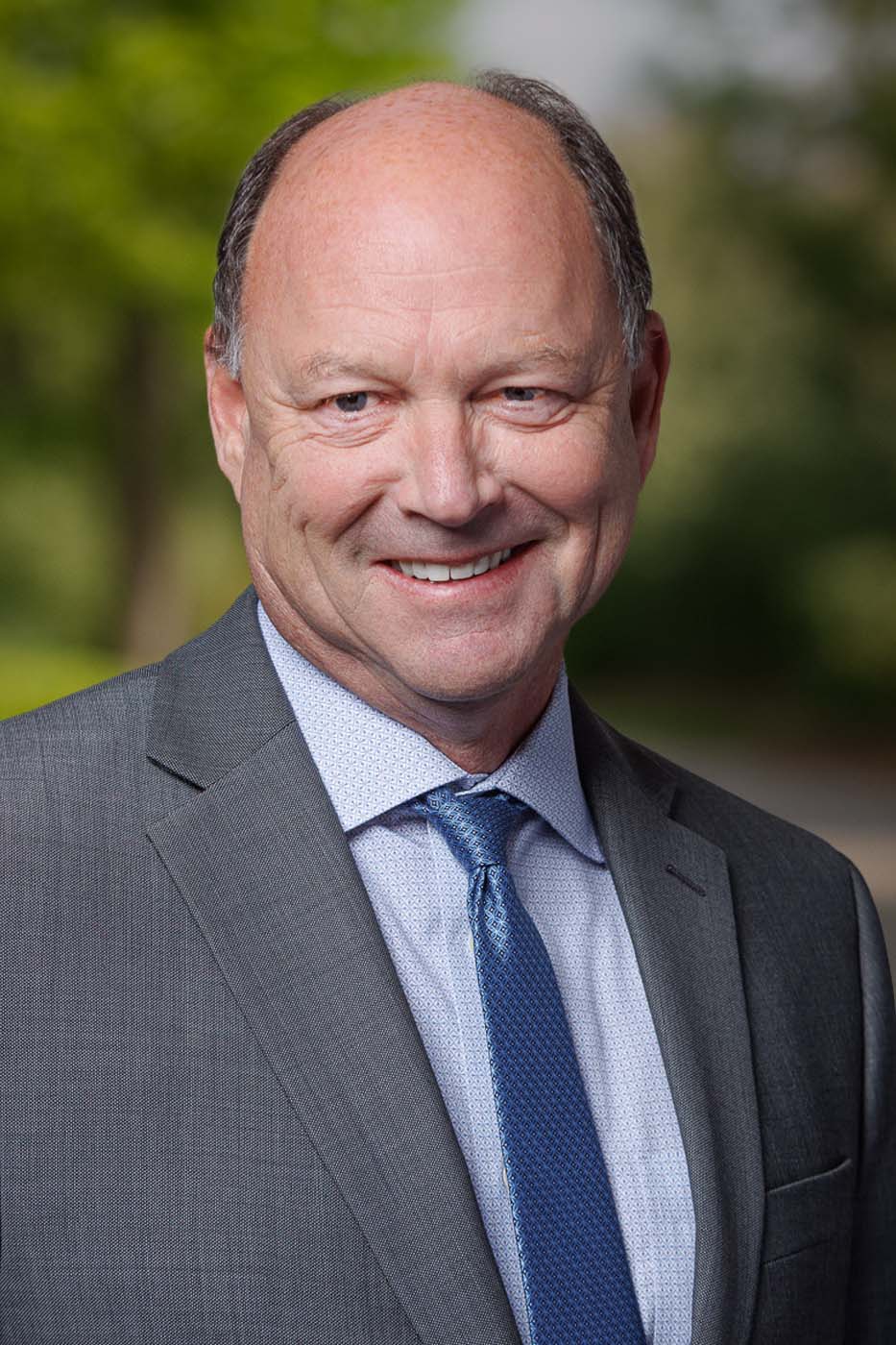 A middle-aged man in a grey suit, light blue shirt, and blue tie is smiling outdoors with a blurred green background, embodying the professionalism of Independent Cost Estimators.