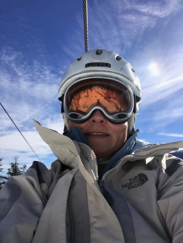 Person wearing a ski helmet, goggles, and a North Face jacket on a ski lift, with blue sky and clouds in the background.