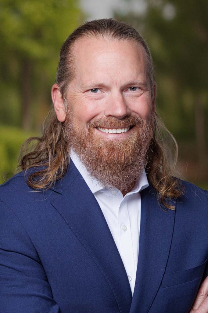 A man with long hair and a beard, wearing a blue suit and white shirt, smiles at the camera with a blurred green outdoor background, embodying the professionalism often seen among Independent Cost Estimators.