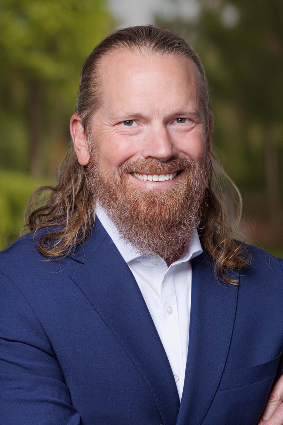 A man with long hair and a beard, wearing a blue suit and white shirt, smiles at the camera with a blurred green outdoor background, embodying the professionalism often seen among Independent Cost Estimators.