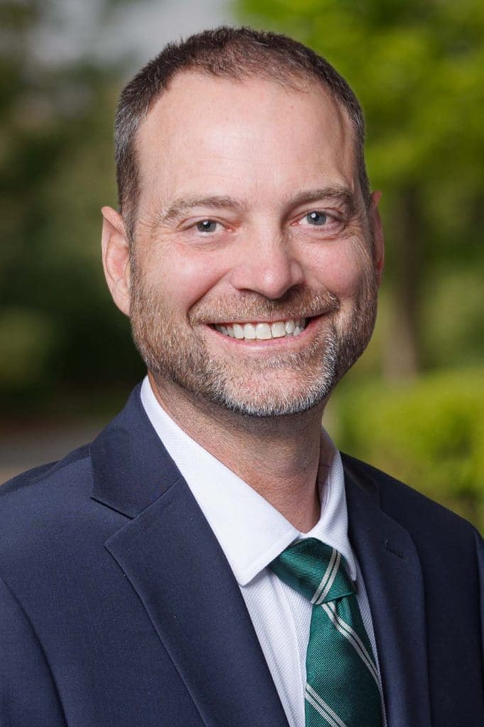 Man in a navy blue suit, white shirt, and green striped tie smiles outdoors with a blurred green background—professional and approachable, ideal for Independent Cost Estimators.