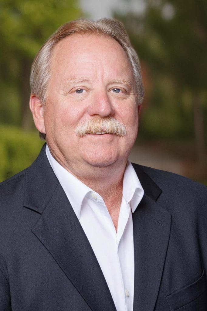 A middle-aged man with light hair and a mustache, looking like one of the Independent Cost Estimators, stands outdoors in a dark suit jacket and white shirt, with greenery in the background.