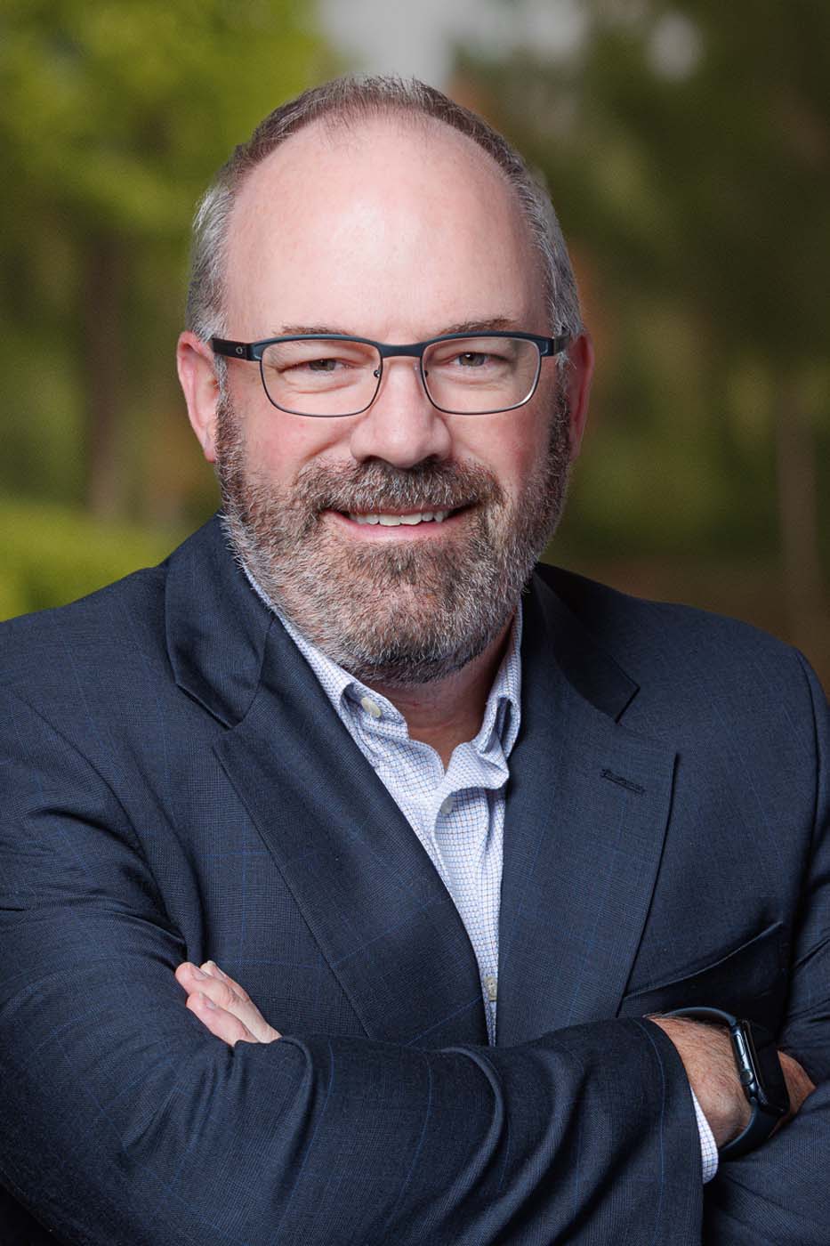 A man with glasses and a beard, resembling an Independent Cost Estimator, stands outdoors in a dark suit jacket and light shirt, arms crossed and smiling confidently at the camera.