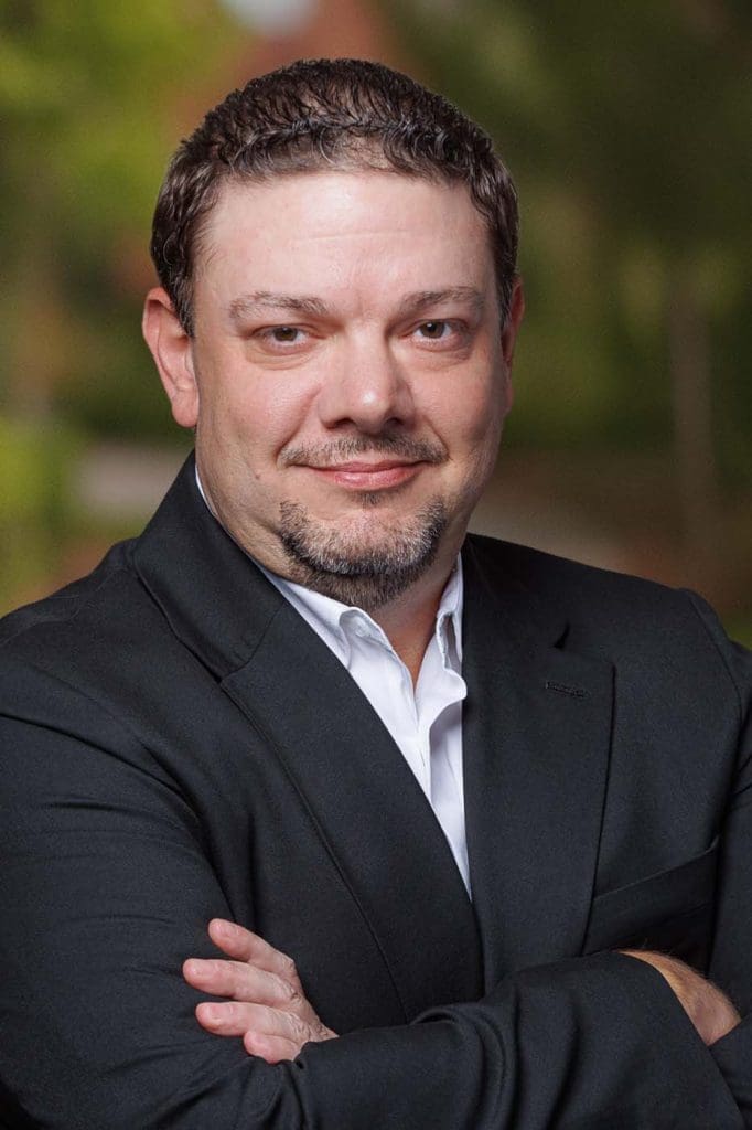 A man with short brown hair and a trimmed beard is wearing a black suit jacket and white shirt, standing with arms crossed and smiling at the camera, embodying the confidence of experienced Independent Cost Estimators.