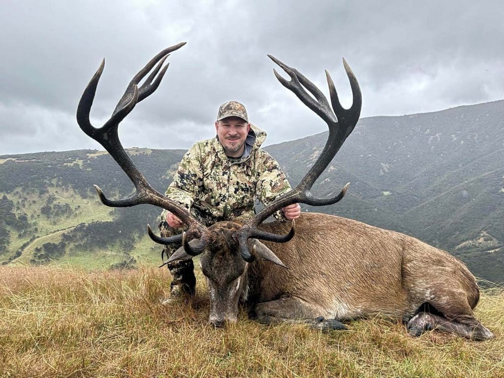 A person in camouflage clothing kneels behind a large deer with antlers on a grassy hillside, with mountains and overcast skies in the background.