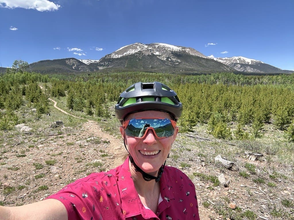A person wearing a bike helmet and sunglasses smiles in front of a forested landscape with snow-capped mountains under a clear blue sky.