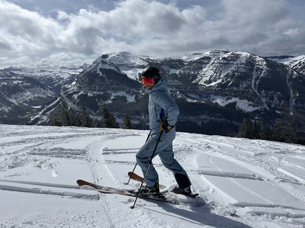 A person in blue ski gear stands on a snow-covered slope with ski poles, mountains and cloudy sky in the background.