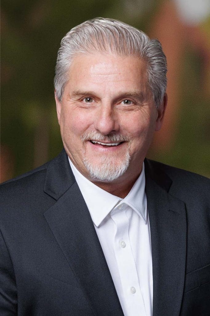 Smiling older man with gray hair and goatee, wearing a dark suit jacket and white shirt, posed in front of a blurred outdoor background—an experienced leader among Independent Cost Estimators.